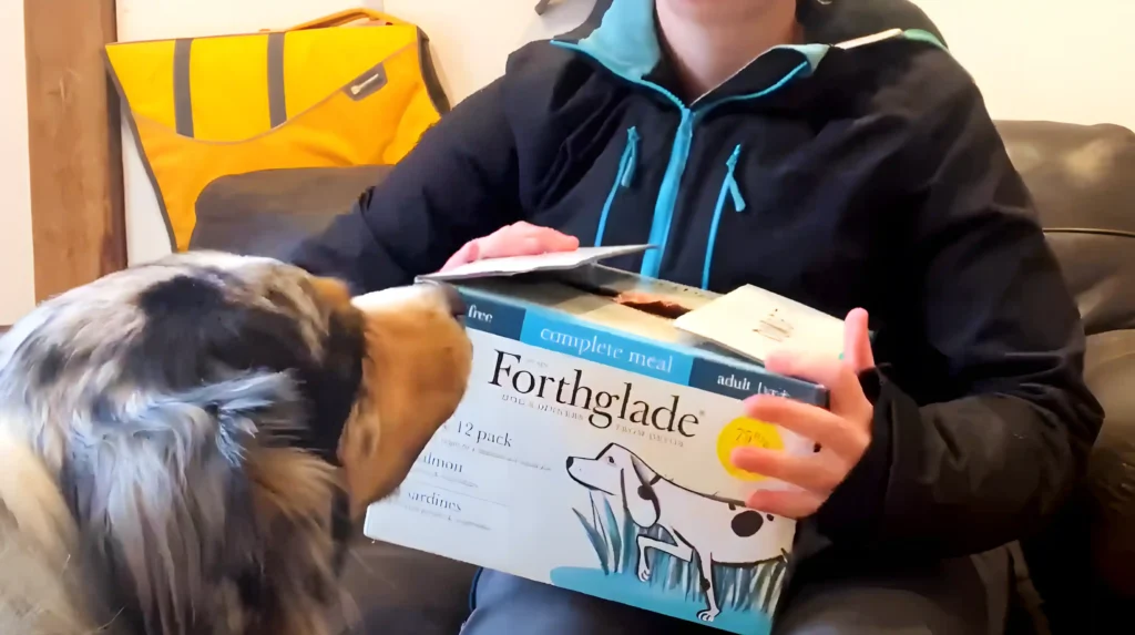 a woman sitting on a couch with a dog hold Forthglade wet dog food box